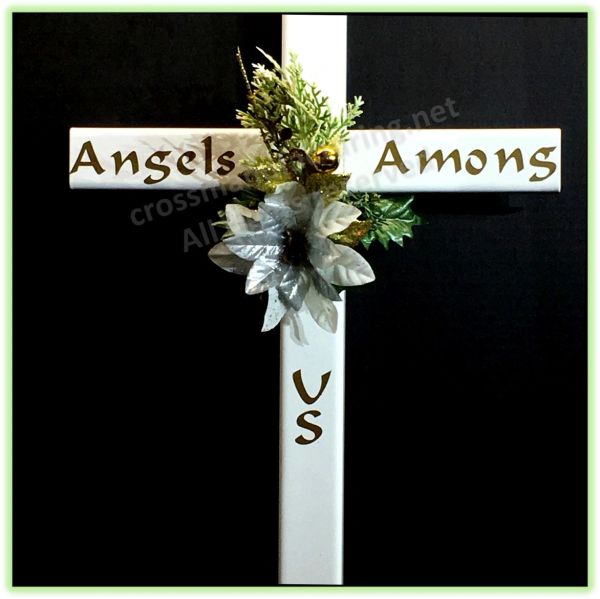 A white STEEL Christian cross with 'Angels Among' written on the horizontal arm and 'US' on the vertical arm, decorated with artificial green foliage and a white flower, shown against a black background.