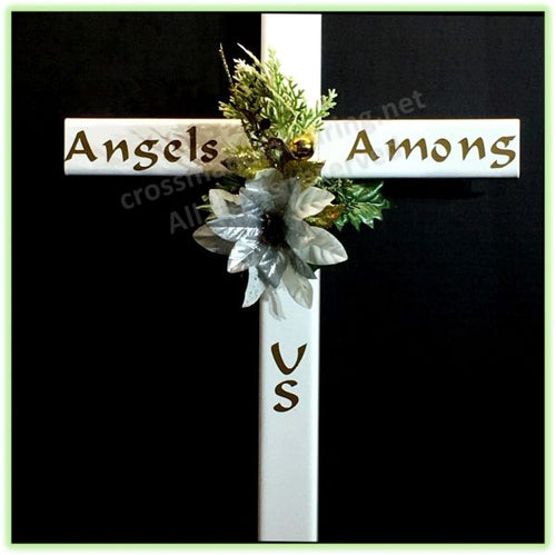 A white STEEL Christian cross with 'Angels Among' written on the horizontal arm and 'US' on the vertical arm, decorated with artificial green foliage and a white flower, shown against a black background.